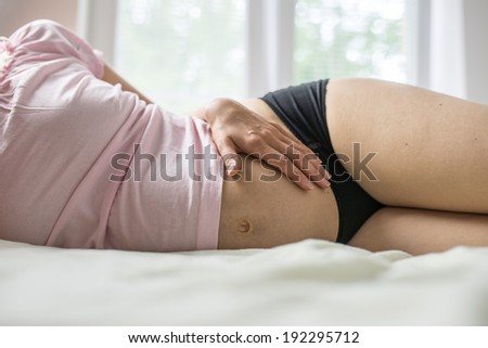 Pregnant woman lying on a bed in panties and a t-shirt holding her swollen belly with her hand as she forms a loving bond with her unborn child, close up torso view.