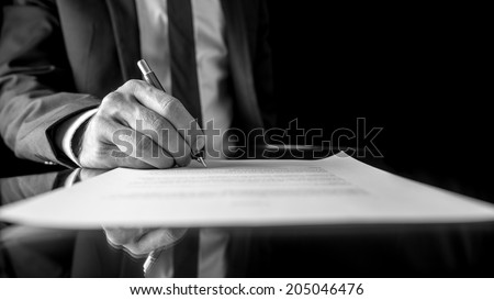 Black and white low angle image of the hand of a businessman in a suit signing a document or contract with a fountain pen on a reflective surface.