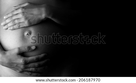 Artistic black and white close-up of an expectant mother holding her belly, concept of care, health and pregnancy, in low-key, with copy space on black.