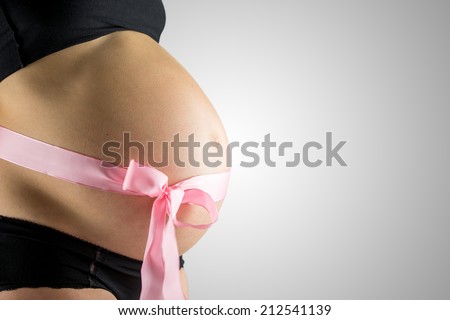 Pregnant woman wearing a decorative pink bow around her bare belly depicting the gender of the unborn baby as being a girl, side view over grey with copyspace.