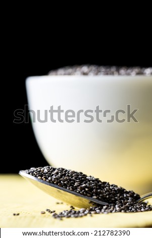 Dried chia seeds from the Salvia hispanica plant rich in omega-3 fatty acids and nutrients, in a spoon overflowing onto a table with a full bowl behind against a dark background.