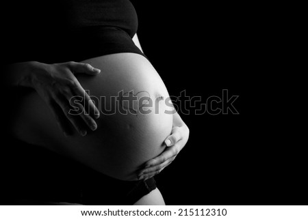 Artistic black and white image of a hand-drawn heart on the bare swollen belly of a pregnant woman cupped by her hands. Isolated over black background.