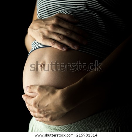 Swollen belly of a pregnant woman cupped by her hands. Over black background.