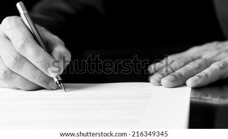 Black and white close up image of the hands of a businessman writing or signing a document with a fountain pen.