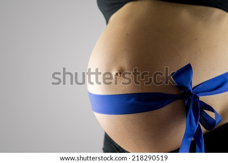 Pregnant woman wearing a blue ribbon around her swollen belly signifying that she is expecting, or hoping for, a baby boy, closeup angled view of her bare belly over grey with copyspace.
