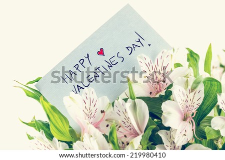 Vintage style Valentines background with a handwritten note saying - Happy Valentines Day - with a small red heart in a bouquet of fresh day lilies on a white background.