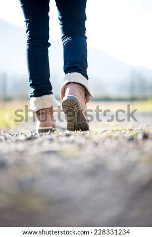 Feet of a woman in jeans and boots walking along a rural path away from the camera.
