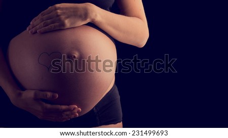 Hand-drawn heart on the bare swollen belly of a pregnant woman cupped by her hands in a concept of bonding an unborn child in a moment of tenderness, vintage effect toned image.