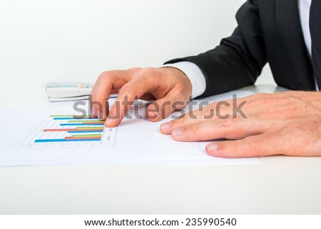 Side view of businessman analysing a set of bar graphs as he sits at his white desk pointing to one column chart, in a business analysis and strategy concept.