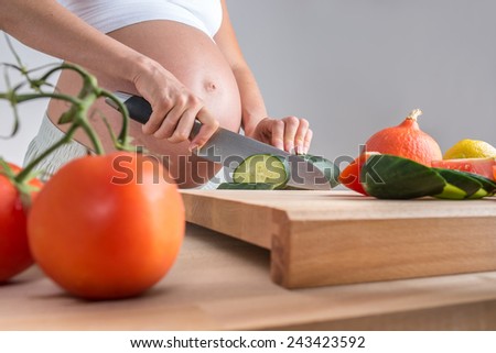 Low angle view of the bare swollen belly of a pregnant woman standing at a kitchen counter with a wooden board chopping vegetables with a knife.