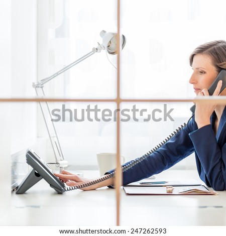 Businesswoman or personal assistant on a telephone call dialing out on a land line instrument as she sits at her desk, side view through an internal window.