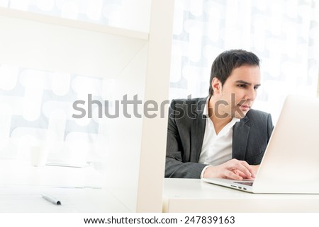 View past an internal partition in a high key office of a hardworking young businessman at his desk reading information on his laptop computer with a serious expression of concentration.