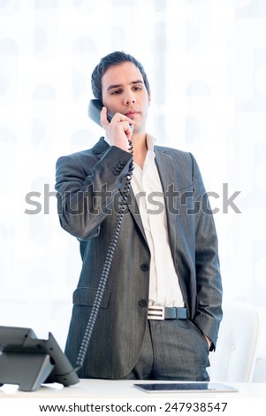 Stylish businessman standing talking on a land line phone in his office listening to the conversation in a relaxed stance with his hand in his pocket.