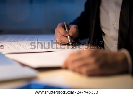 Businessman working late signing a document or contract in a dark office with a fountain pen by the light of a lamp, close up view of his hands.