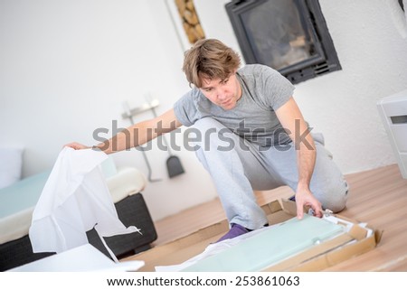 Man in casual clothes kneeling unpacking a package on the wooden floor in the living room at home with an insert wood burner fire in the wall behind him.