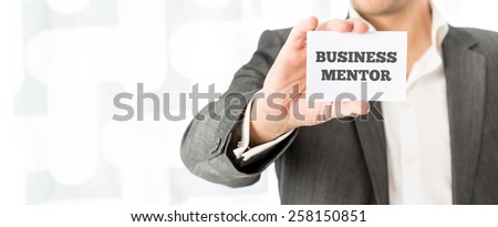 Businessman holding up a small white card with the words Business Mentor in a concept of consulting, advice, skill and experience.