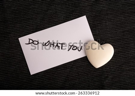 White card with a Do what you love sign with a ceramic heart instead of the word love. Card and heart placed on a dark blue ribbed textile.