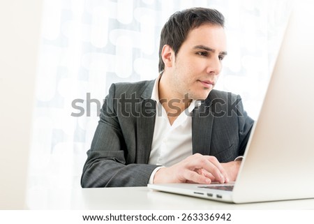 Portrait of a businessman in a high key office typing information on a white laptop computer keyboard, low angle view from the back of the computer.