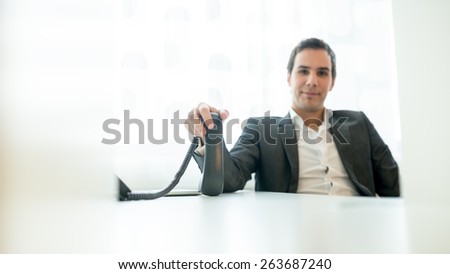 Young Businessman Sitting at his Worktable Holding a Telephone While Looking at the Camera.