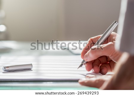 View past the arm of a colleague of a businessman writing on a document with a fountain pen, close up focus to the hand.