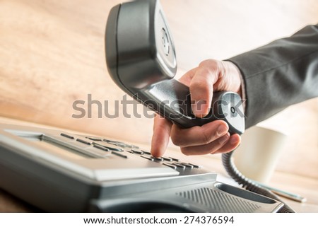 Hand of a businessman with dark gray suit holding the receiver of a black corded desk phone while dialing, in the office.