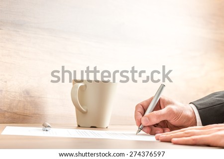 Hands of a businessman writing with a silver pen on a paper sheet, at a wooden desk, next to a mug of coffee or cappuccino, with copy space on blurred beige wall.