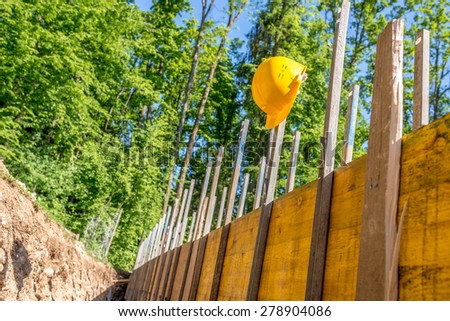 Bright Yellow Hard Hat Hanging on Post of Unfinished Building Foundation Under Construction in Wooded Forest Area.