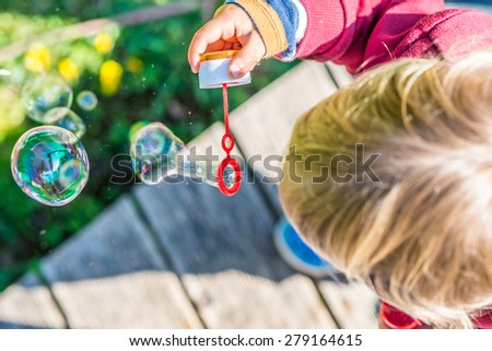 Overhead view of the top of the head of a young blond child blowing colorful iridescent bubbles with a soap solution while standing outdoors on a wooden deck in the garden.