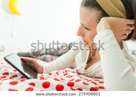 Young woman wearing a golden alice band in her hair looking on digital tablet or e-book device lying on her bed on a red and white polka dot bedspread.