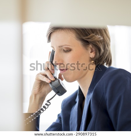 View through an internal office partition of a successful young businesswoman sitting at her desk making a phone call on a landline telephone, profile view.