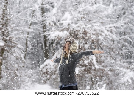 Happy young woman embracing beautiful life with her arms wide open as she is standing outside in a snowy winter woodland with snowflakes falling.