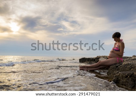 Pregnant woman in ninth month of pregnancy sitting on a rock by the sea cradling her swollen belly as she bonds with her unborn child.