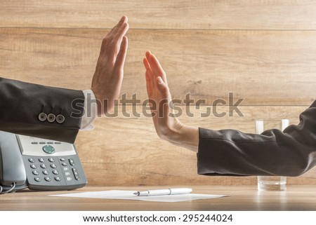 Two hands reach to hi-five across business table in order to celebrate a successful business deal, arms in black suits, telephone, document with pen and glass of water visible, wooden background.