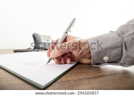 Closeup of businessman or lawyer signing an important contract, document or paperwork on a wooden desk with a landline telephone in background.