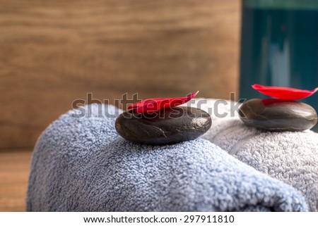 Luxurious spa arrangement for two with a a blue and white folded soft towel, black massage stones with red rose petal on top and a bottle of massage oil or moisturizing lotion.