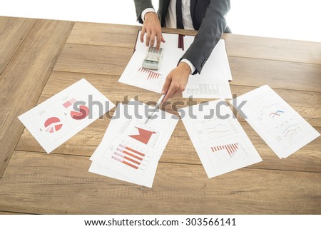 Businessman giving a presentation with assorted analytical graphs and charts spread out on the desk in front of him pointing to relevant information with a pen.