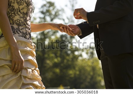 Man proposing or marrying his sweetheart outdoors in a park with a low angle view looking up of him about to place a ring on the finger of her left hand, close up torso view.