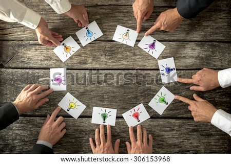 Teamwork and brainstorming concept with businessmen seated around a table each pointing to cards with colorful sketches of light bulbs conceptual of bright ideas and solutions arranged in a circle.
