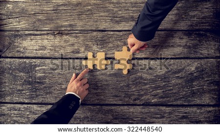 Retro style image of two business partners each placing one matching piece of puzzle on a textured wooden table. Conceptual of cooperation, innovation and success.
