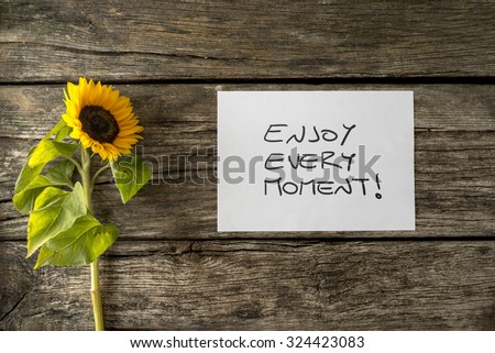 White card with an Enjoy every moment message lying next to a beautiful blooming sunflower on a textured wooden desk in order to encourage you to seize the moment and live a fulfilled life.