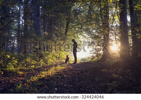 Man walking his dog in the woods standing backlit by the rising sun casting a warm glow and long shadows.