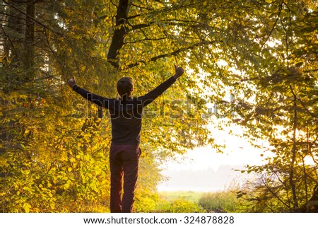Young man standing on the edge of forested area raising his arms in celebration turned towards a bright sunlight waiting for him just outside the forest. Concept of freedom, achievement and victory.