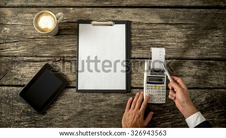 Business accountant or financial adviser checking income and expenses in order to write an annual report as he makes calculations on adding machine. With blank sheet of paper in front of him.