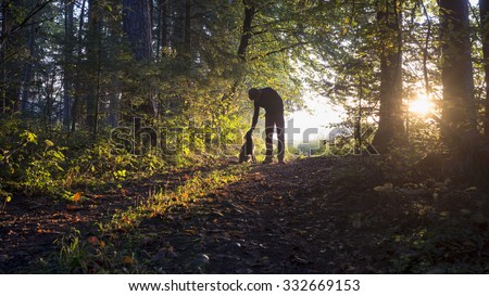 Man bending down to pet his black dog as they enjoy a beautiful nature in a glade in the woods backlit by the warm glow of the early morning sun.