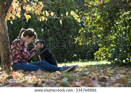 Young woman sitting under a colorful autumn tree lovingly petting her black dog as they join their noses in affection.