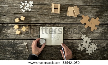 Businessman making plan and business strategy decisions as he sketches a compass he is holding into his notebook. Various cubes, pegs, puzzles and a key lying on his wooden office desk, top view.