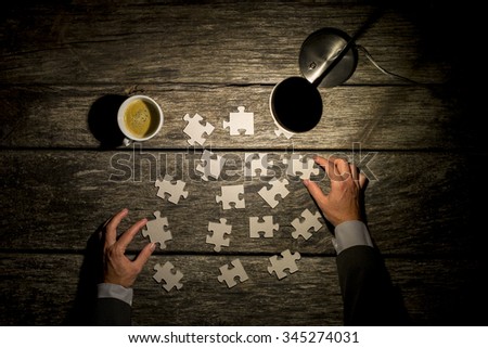 Top view of male hands in elegant business suit trying to find a solution to a problem by arranging and matching puzzle pieces on a textured rustic wooden desk wit a lit table lamp and cup of coffee.