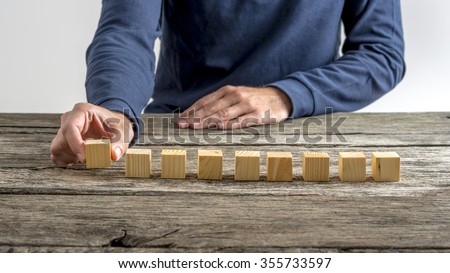 Front view of male hand placing nine blank wooden cubes in a row on a textured rustic wooden desk.