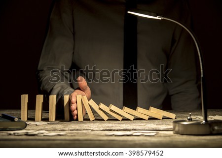 Front view of businessman stopping falling dominos with his hand on wooden textured office desk with a lamp turned on.