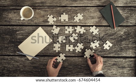 Retro style image of male hands in business suit trying to find a solution to a problem by arranging and matching puzzle pieces on a textured rustic wooden desk, top view.